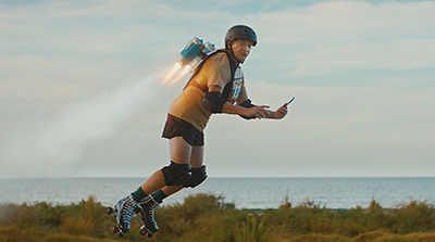  A person wearing a helmet, knee pads, and roller skates is propelled forward by a jetpack, with visible flames and smoke coming from the pack. The individual is holding a smartphone while skating outdoors near greenery, with a body of water and sky in the background.