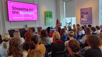 A speaker addresses an audience seated in a bright conference room during an event. A large screen displays the text “Stepping into the Spotlight – International Women’s Day Special,” and colorful posters with text are visible on the walls, including one that reads “Pick & Mix.”